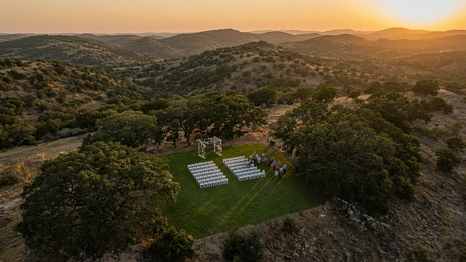 Aerial view at sunset of an outdoor ceremony with rows of white chairs on a Hill Country ranch lawn